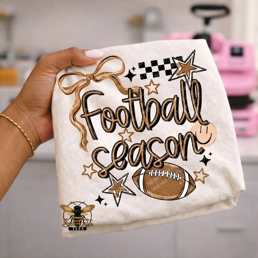 A hand holds a folded white fabric item featuring a gold and black 'Football Season' graphic with stars, a football, and a bee logo. The background is a softly blurred kitchen setting with a pink appliance visible.