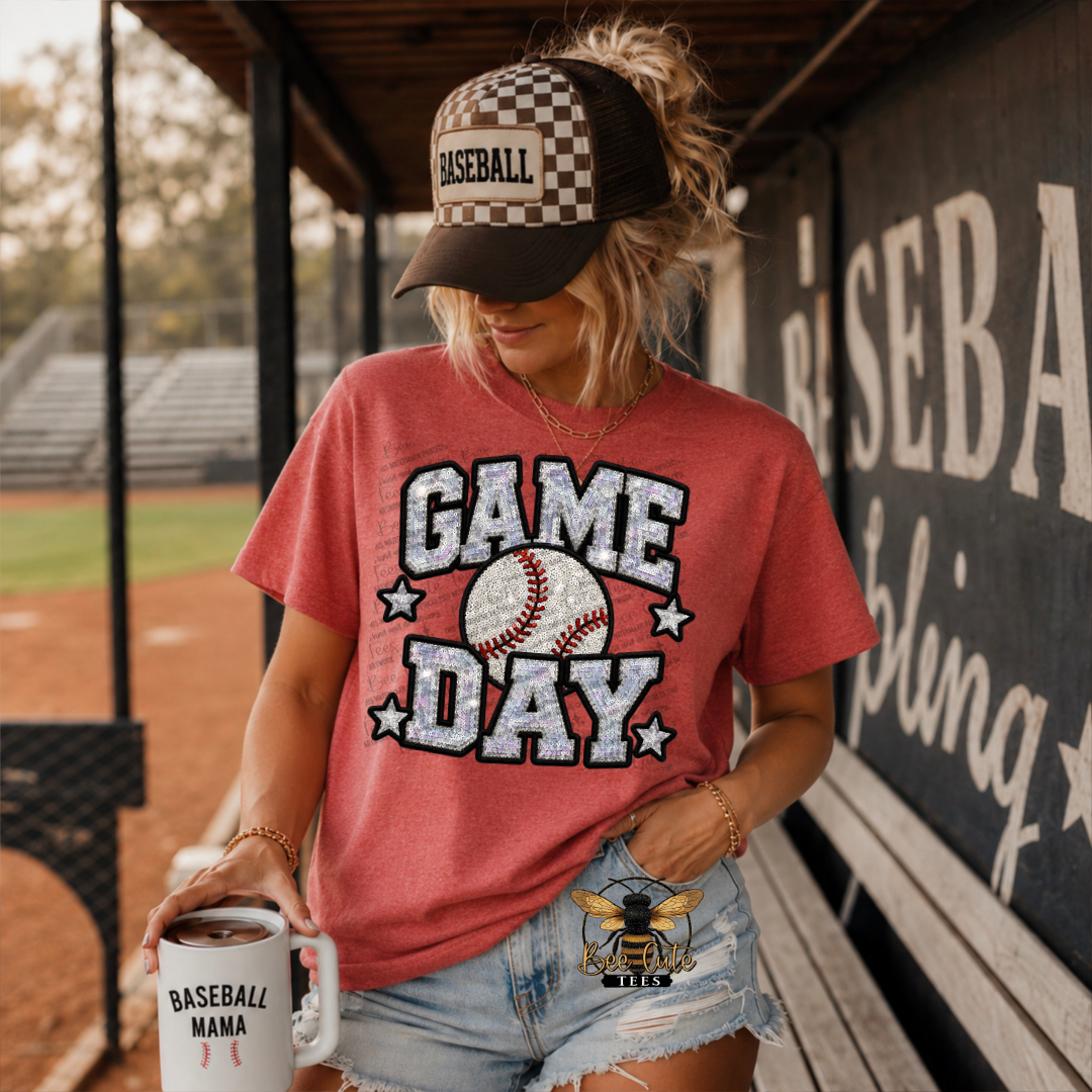 A woman stands in a baseball dugout wearing a coral-red graphic T-shirt that reads 'GAME DAY' with a baseball and stars, paired with denim shorts and a checkered baseball cap. She holds a white mug that says 'BASEBALL MAMA'.