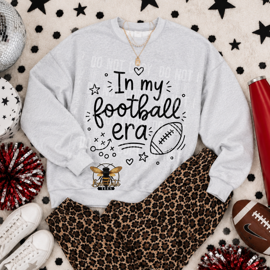A light gray crewneck sweatshirt with a 'In my football era' graphic, styled in a flat lay with leopard print pants, white sneakers, a red tumbler, and sports-themed props on a patterned rug.
