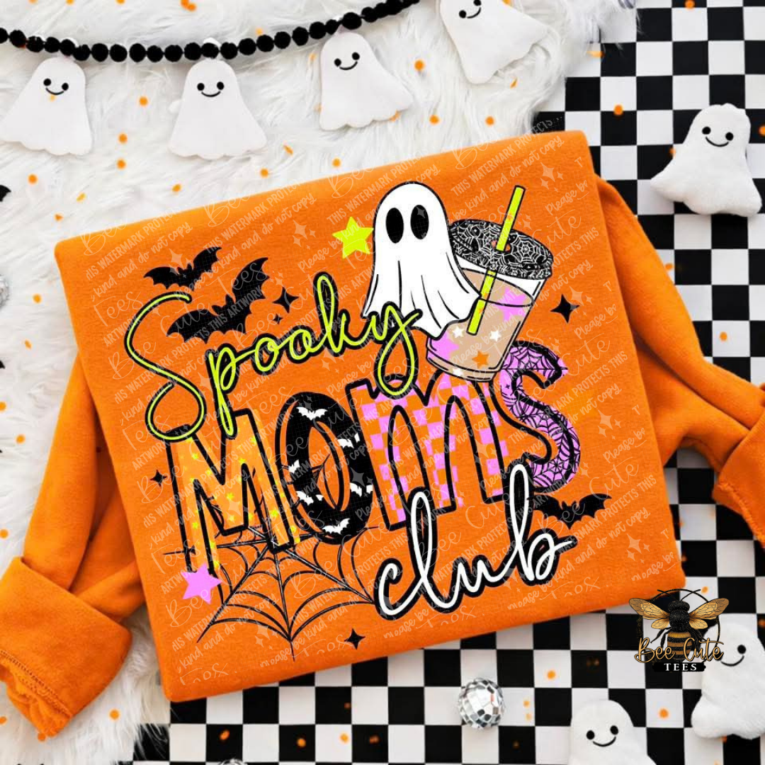 An orange long-sleeve sweatshirt with a 'Spooky Moms Club' graphic design, styled in a flat-lay arrangement with Halloween props including ghosts, a black and white checkered mat, and a black beaded garland.