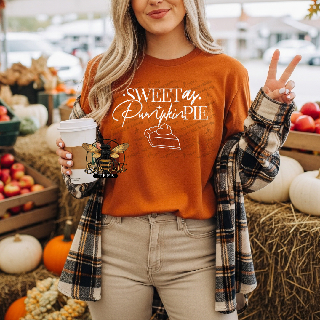 A woman wearing an orange crewneck sweatshirt with a 'Sweet as Pumpkin Pie' graphic, styled with a plaid flannel shirt and beige pants, standing in a fall-themed outdoor market setting.