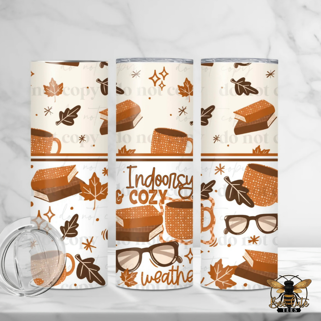 Three insulated tumblers with a fall-themed pattern featuring books, coffee cups, autumn leaves, glasses, and the words 'Indoors cozy weather' on a white marble background. A clear lid is shown in the lower left.