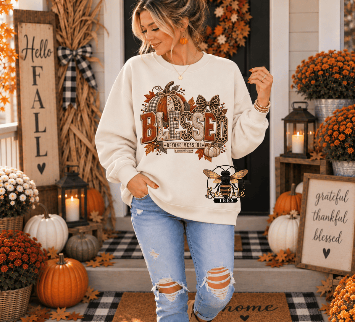 A woman stands on a decorated front porch wearing a cream sweatshirt with a large autumn-themed 'Blessed' graphic and distressed light-wash jeans. The scene includes pumpkins, mums, lanterns, fall foliage, and seasonal decor.