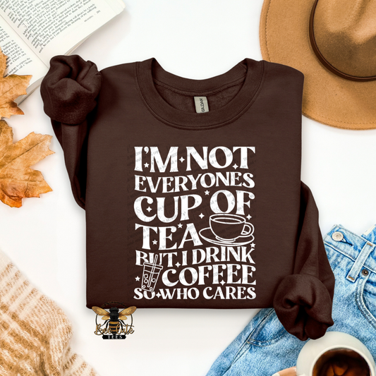 A dark brown crewneck sweatshirt with a white graphic text print reading 'I'M NOT EVERYONE'S CUP OF TEA BUT I DRINK COFFEE SO WHO CARES' with a coffee cup illustration. The sweatshirt is styled in a flat-lay arrangement with a tan hat, blue jeans, a book, autumn leaves, a knit sweater, and a cup of coffee on a white background.