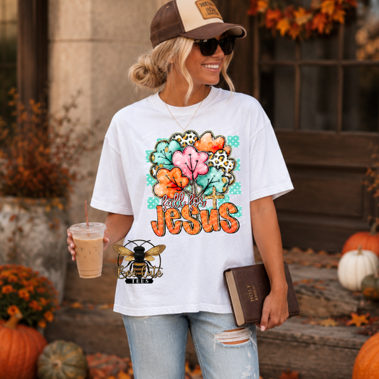 A woman wearing a white graphic t-shirt with a colorful floral and cross design that reads 'FALL FOR JESUS' in orange, standing outdoors in front of a rustic door with pumpkins and autumn decorations.