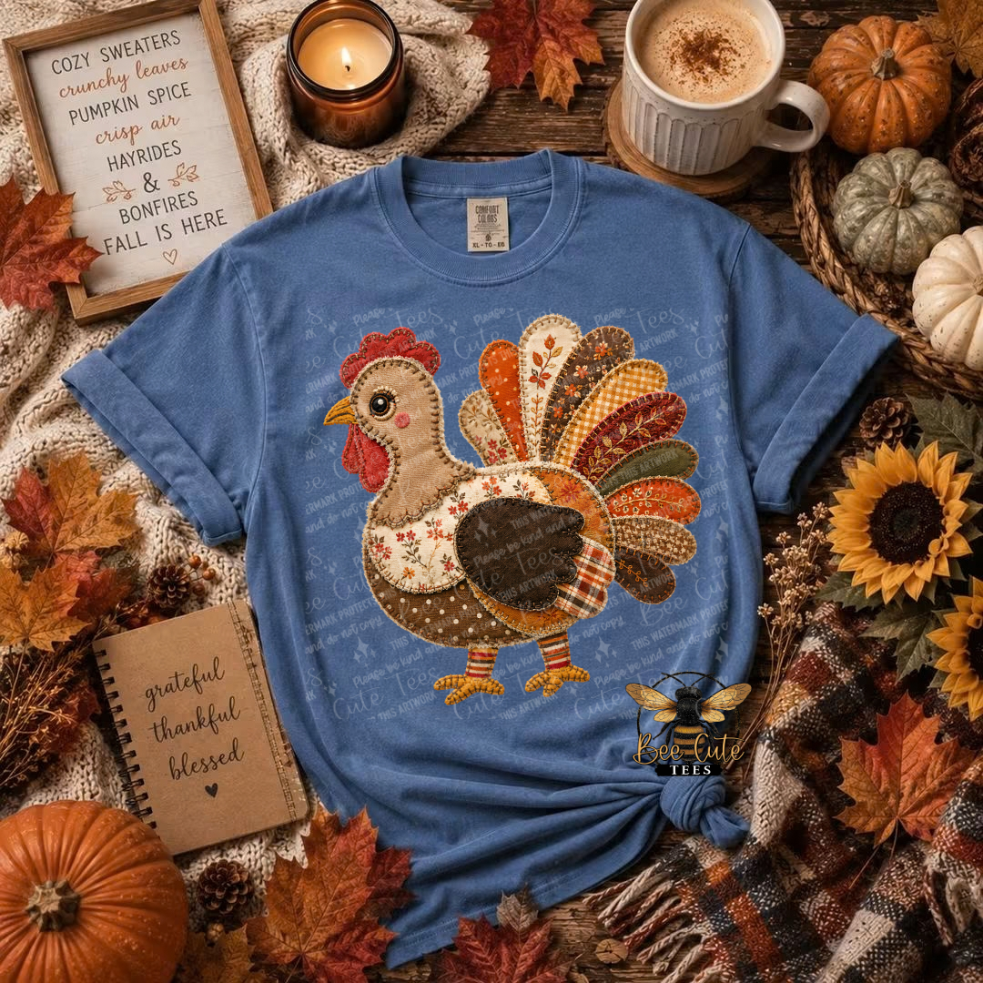 A blue graphic t-shirt featuring a patchwork-style turkey design in autumn colors, styled in a cozy fall flat-lay with pumpkins, sunflowers, a mug of coffee, and autumn leaves.