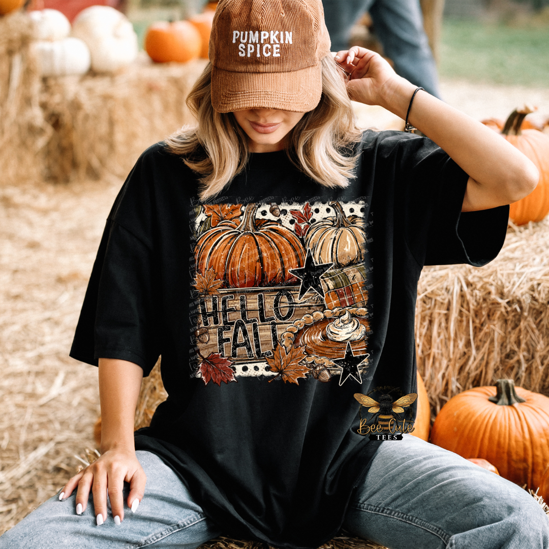A woman sitting outdoors on hay bales wears a black graphic T-shirt with a fall-themed pumpkin and 'HELLO FALL' print, along with a brown 'PUMPKIN SPICE' cap. Pumpkins and hay are visible in the background.