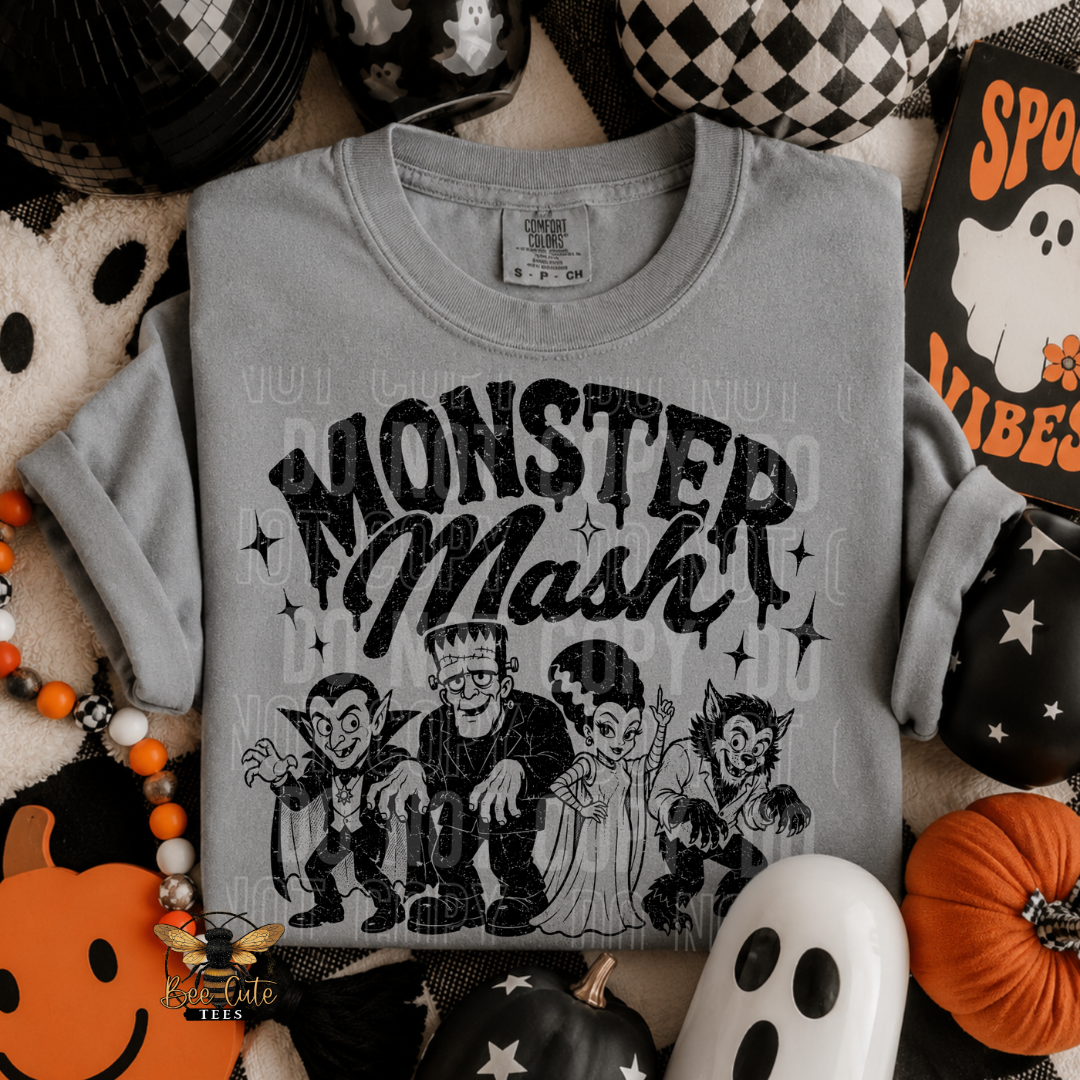 A light gray short-sleeve t-shirt with a black 'Monster Mash' graphic featuring Frankenstein, Dracula, Vampire, and Wednesday Addams, styled in a flat-lay Halloween arrangement with pumpkins, a ghost mouse, and themed props.
