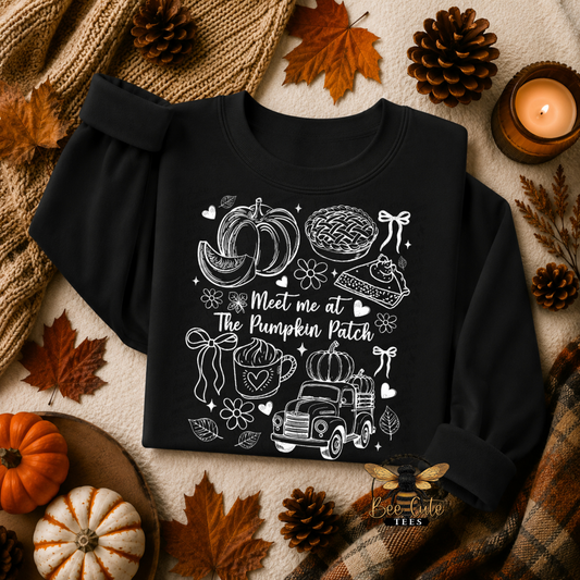 A black long-sleeve crewneck sweatshirt with a white autumn-themed graphic that reads 'Meet me at The Pumpkin Patch' and includes pumpkins, a pie, a mug, flowers, and a truck. The sweatshirt is styled in a flat lay with fall leaves, pinecones, a candle, and a plaid scarf.