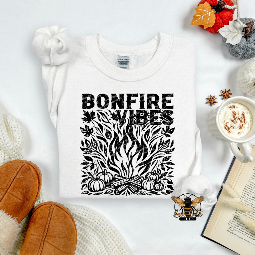A white crewneck sweatshirt with a black graphic reading 'BONFIRE VIBES' above a stylized campfire illustration with pumpkins and leaves. The sweatshirt is presented in a cozy flat-lay arrangement with a knit hat, tan slipper edges, a mug of hot cocoa, a book, and autumn leaves on a white background.