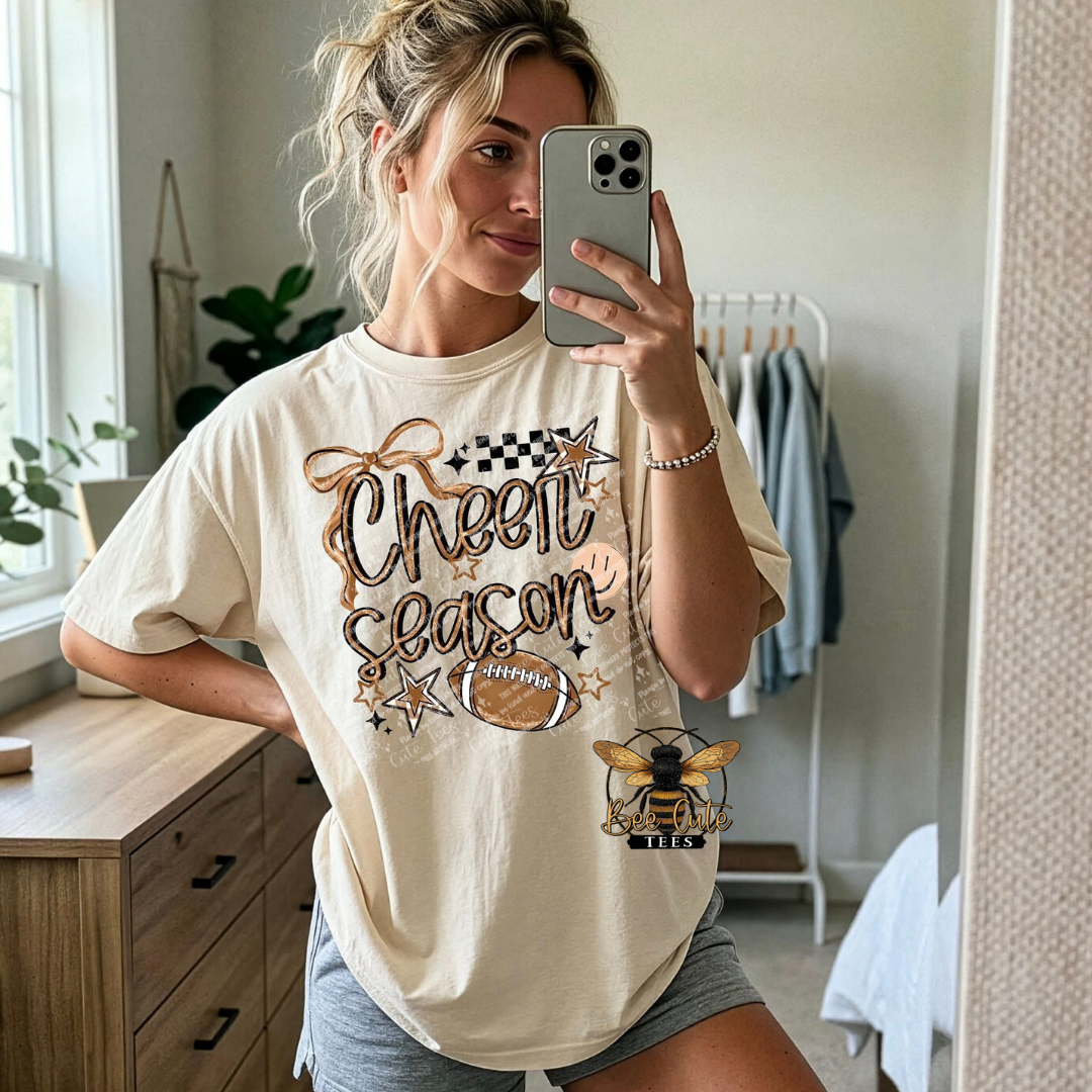 A woman takes a mirror selfie wearing a cream-colored graphic t-shirt with 'Cheer Season' in brown glitter-style lettering, a football graphic, and a bee logo.