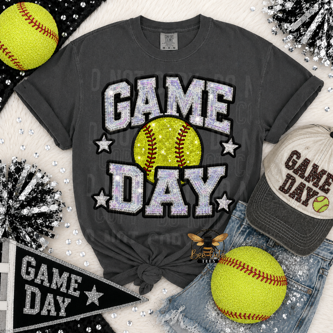 A dark charcoal gray graphic t-shirt featuring 'GAME DAY' in glittery silver letters with a yellow softball in the center. The t-shirt is styled in a flat-lay arrangement with a baseball cap, denim shorts, two yellow softballs, and black-and-silver pom-poms on a white textured background.