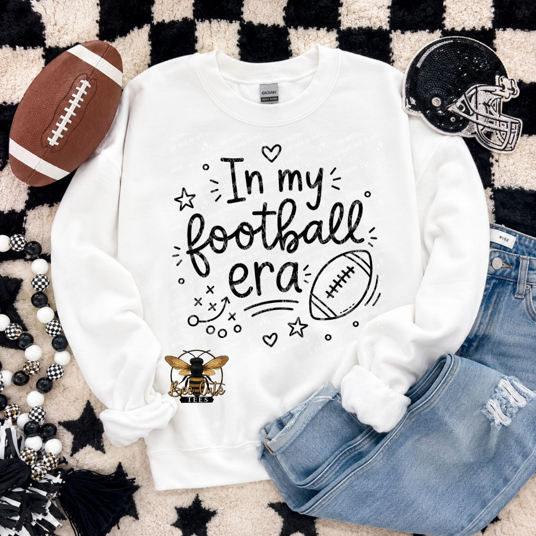 A white crewneck sweatshirt laid flat on a black-and-white checkered rug, surrounded by football-themed props including a brown football, a black helmet, blue jeans, and decorative beads. The sweatshirt has black text reading 'In my football era' with small heart, star, and doodle accents, plus a small bee graphic near the lower left.