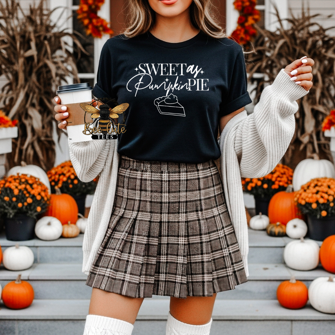 A woman wearing a black graphic t-shirt that reads 'Sweet as Pumpkin Pie' with a slice of pie illustration, styled with a beige knit cardigan and a brown plaid pleated skirt, standing on steps decorated with pumpkins and orange flowers.