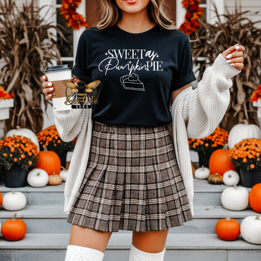 A woman wearing a black graphic t-shirt that reads 'Sweet as Pumpkin Pie' with a slice of pie illustration, styled with a beige knit cardigan and a brown plaid pleated skirt, standing on steps decorated with pumpkins and orange flowers.