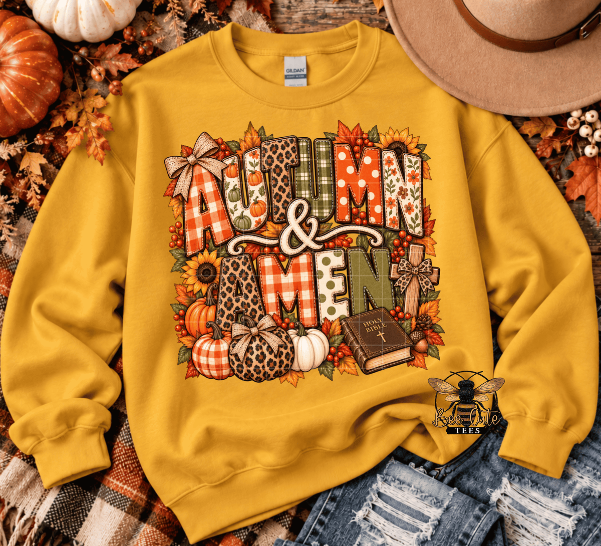 A yellow crewneck sweatshirt with a large 'Autumn & Amen' graphic featuring fall-themed patterns, styled in a flat-lay with pumpkins, a hat, and denim.