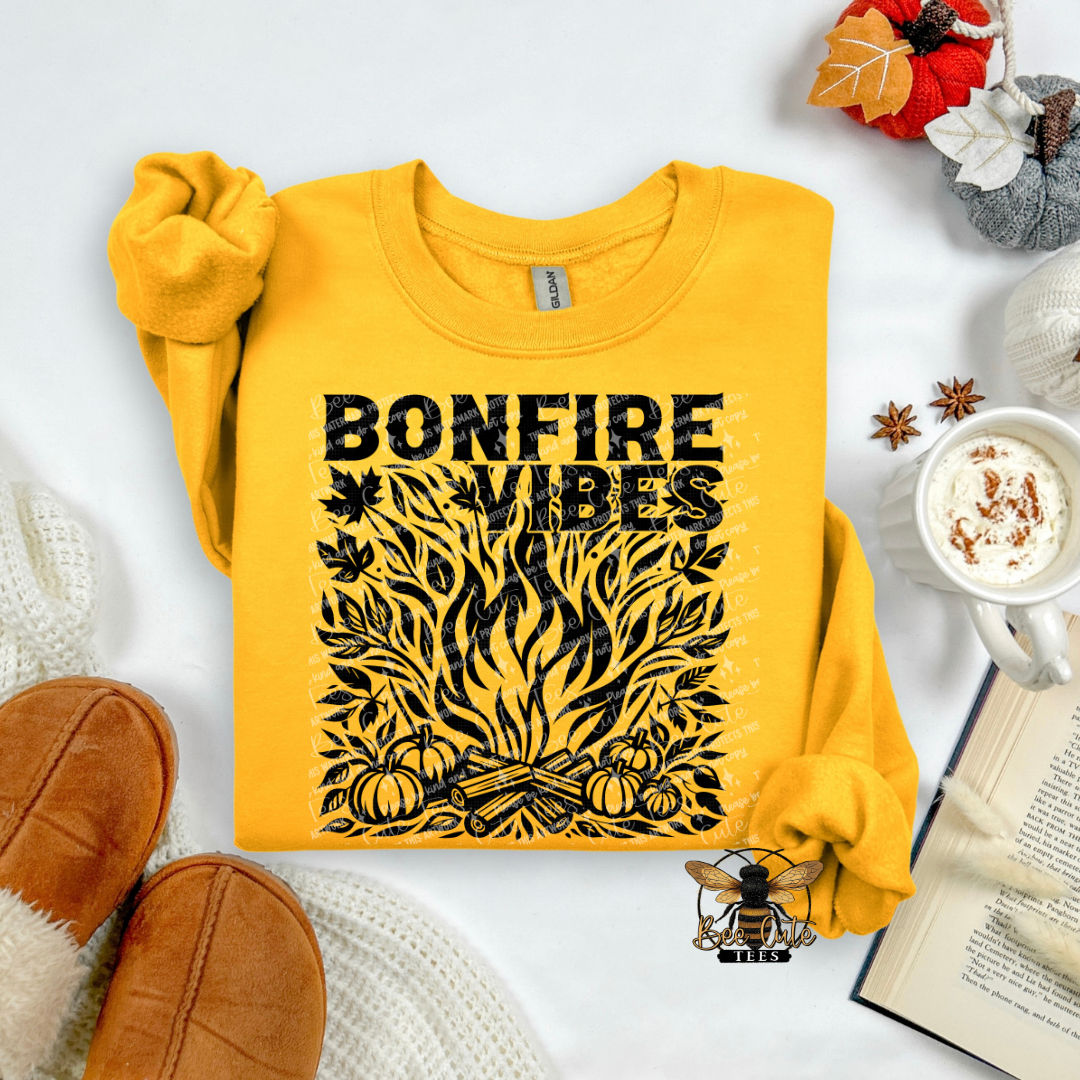 A yellow long-sleeve sweatshirt laid flat on a white surface, featuring a black graphic that reads 'BONFIRE VIBES' above a stylized campfire, pumpkins, leaves, and autumn-themed props including a mug, book, slippers, and decorative pumpkins.