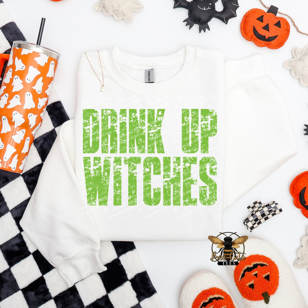 A white crewneck sweatshirt with the phrase 'DRINK UP WITCHES' printed in distressed green letters, styled in a flat lay with Halloween props including a ghost-patterned tumbler, jack-o'-lanterns, and a black-and-white checkered blanket.