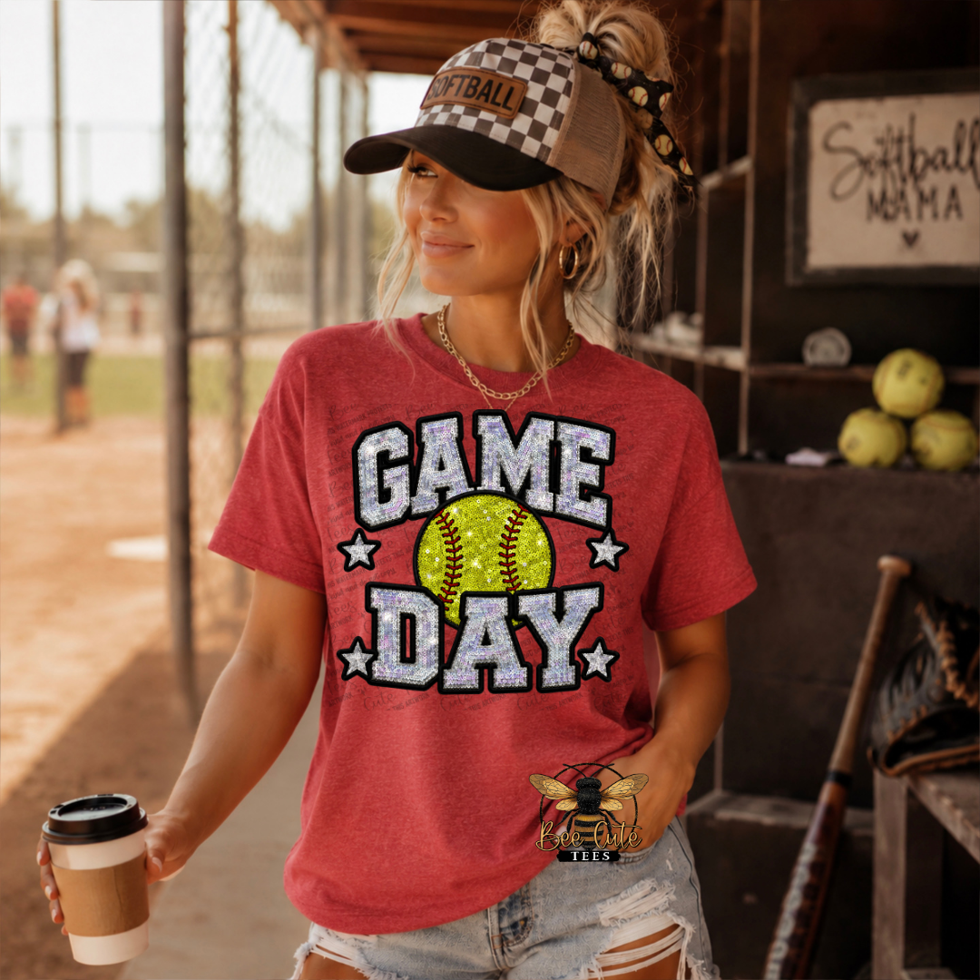 A woman wearing a red graphic t-shirt that says 'GAME DAY' with a softball graphic, a checkered baseball cap, and denim shorts, standing in a softball field setting.