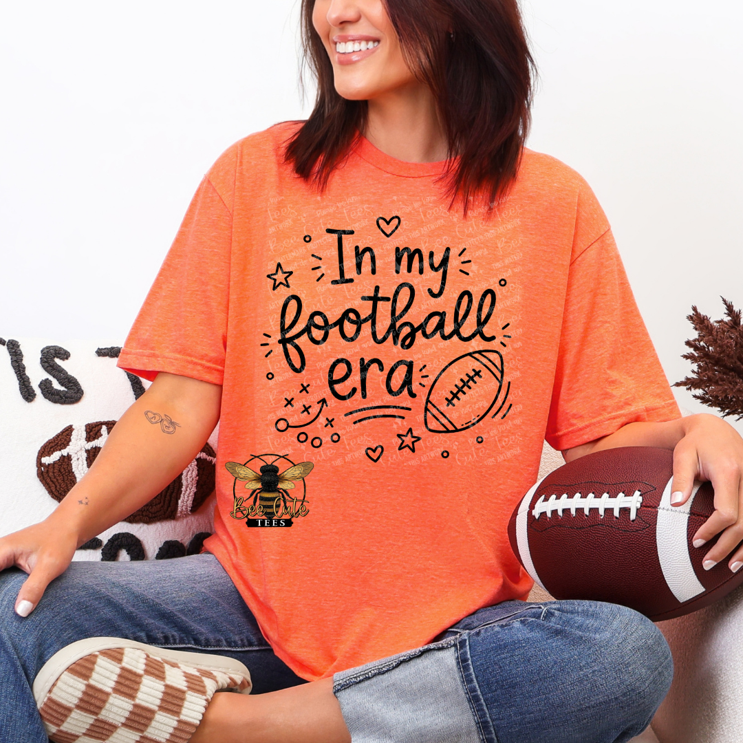 A woman wearing a bright orange graphic t-shirt that reads 'In my football era' with a bee and football design, holding a brown football while sitting.