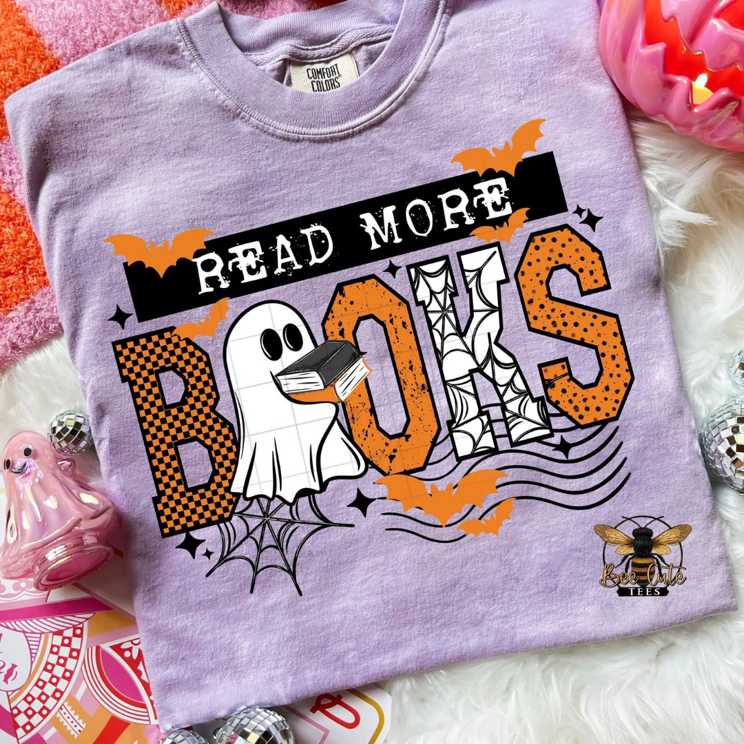 A lavender short-sleeve graphic T-shirt laid flat on a white faux-fur surface with Halloween-themed props. The shirt reads 'READ MORE BOOKS' with a ghost, spiderwebs, bats, and a bee logo on the front.