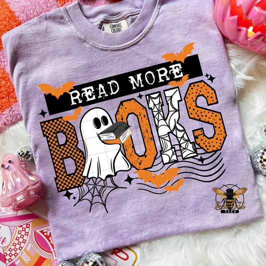 A lavender short-sleeve graphic T-shirt laid flat on a white faux-fur surface with Halloween-themed props. The shirt reads 'READ MORE BOOKS' with a ghost, spiderwebs, bats, and a bee logo on the front.