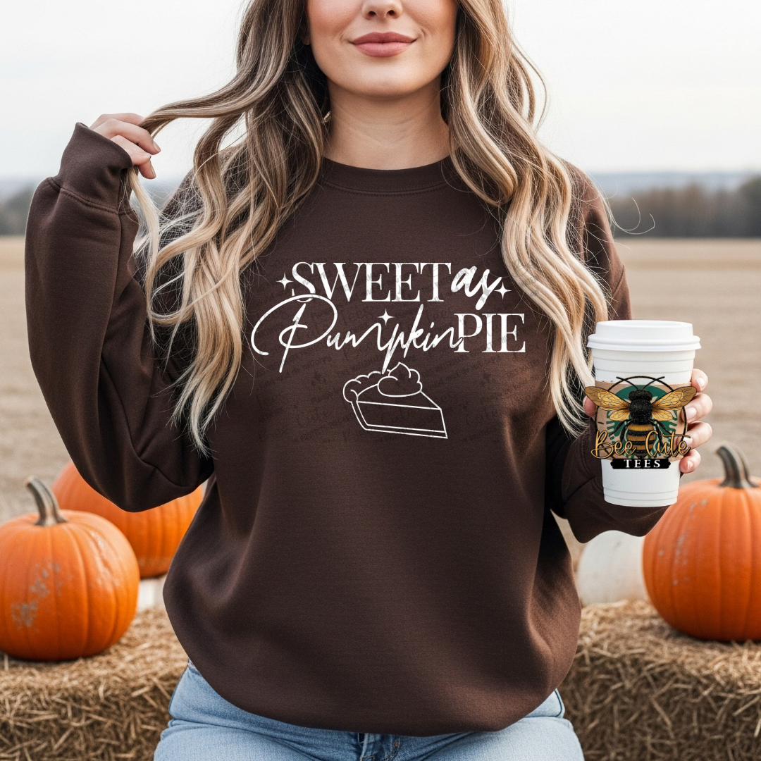 A woman wearing a brown crewneck sweatshirt with a 'Sweet as Pumpkin Pie' graphic, holding a coffee cup in a pumpkin field.