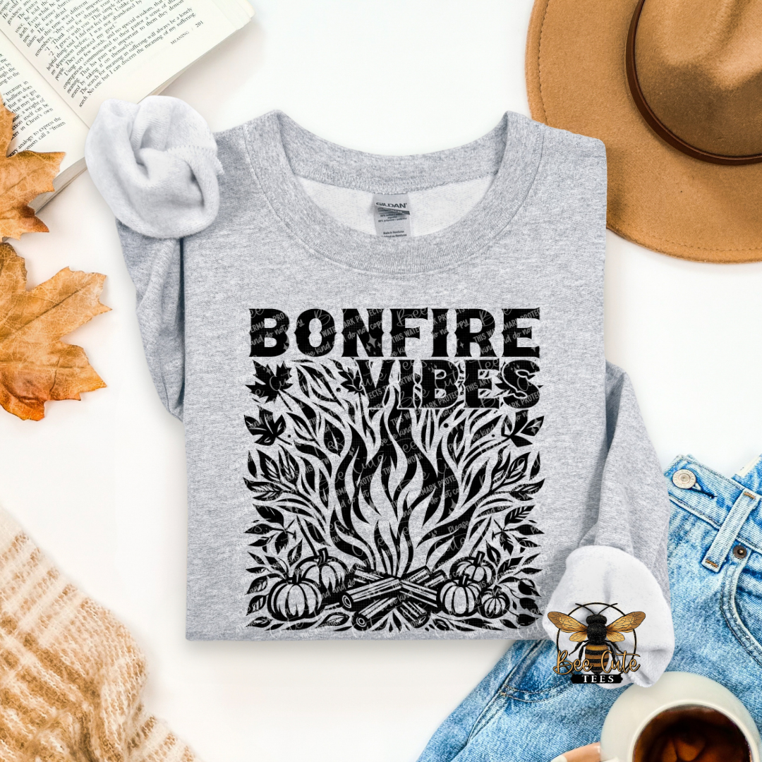 A heather gray long-sleeve t-shirt with a black graphic reading 'BONFIRE VIBES' above a campfire illustration, styled in a flat lay with a hat, jeans, autumn leaves, an open book, a cup of coffee, and a knit sweater on a white background.
