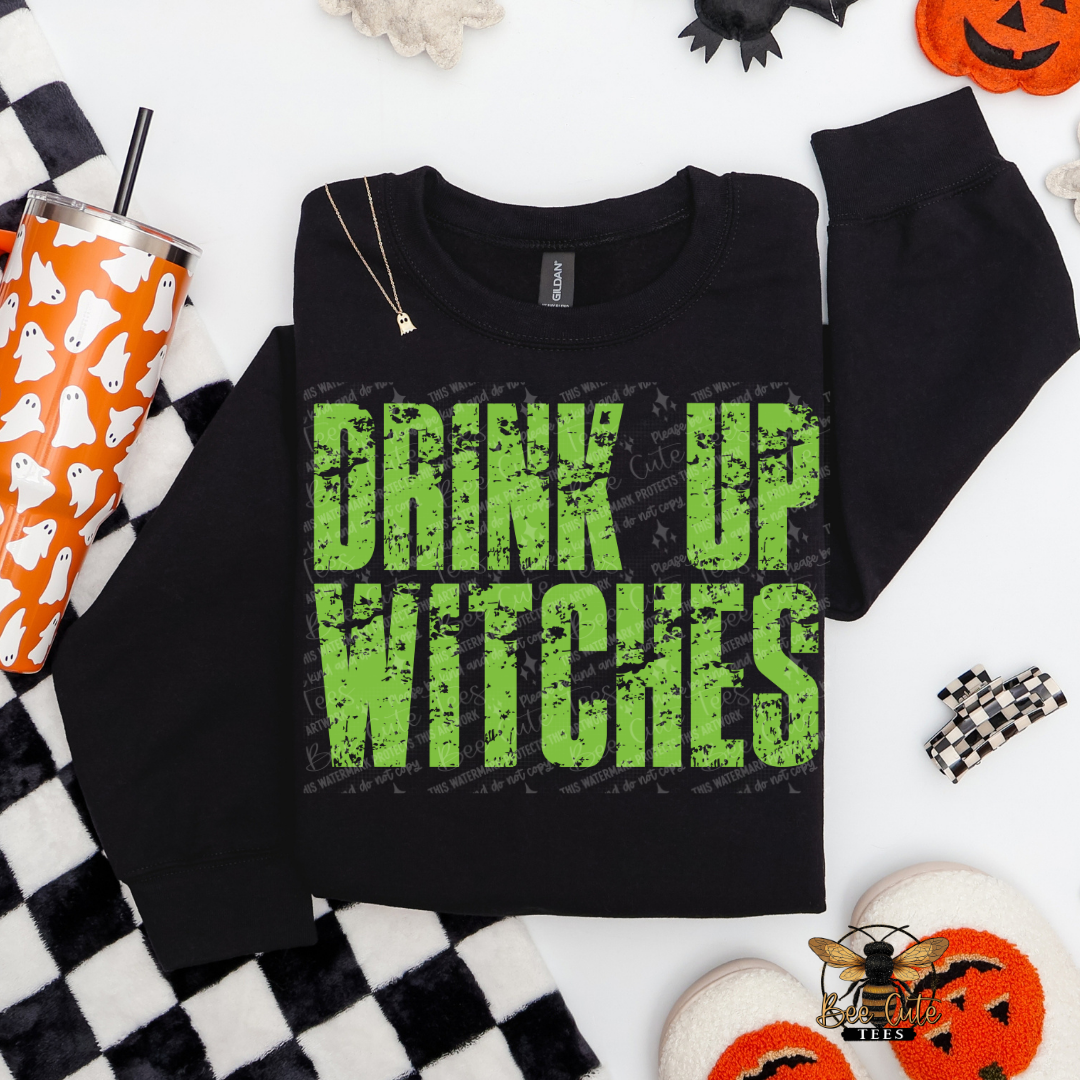 A black crewneck sweatshirt with the phrase 'DRINK UP WITCHES' printed in distressed green letters, styled in a flat lay with Halloween-themed props including a ghost-patterned tumbler, black and white checkered blanket, and orange jack-o'-lantern decorations.