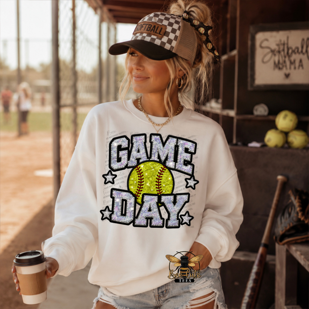 A woman on a softball field stands wearing a white sweatshirt with a glittery 'GAME DAY' graphic and a softball in the center, paired with a checkered baseball cap and denim shorts. She holds a coffee cup in a sunny outdoor sports setting.