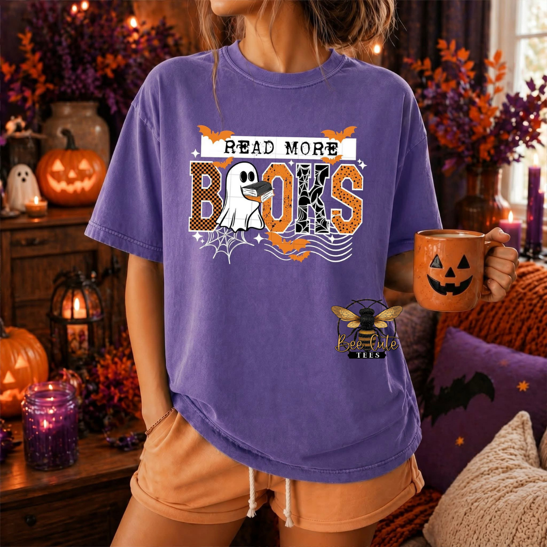 A woman wearing a purple graphic t-shirt with a Halloween design that reads 'READ MORE BOOS' with a ghost, spider, bat, and orange batwing motifs. She is holding an orange jack-o'-lantern mug in a cozy, decorated Halloween-themed room.