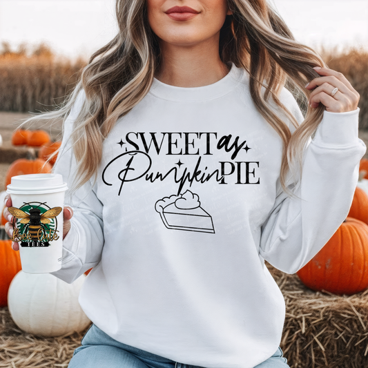 A woman wearing a white sweatshirt with a black graphic that reads 'Sweet as Pumpkin Pie' and a slice of pie illustration, standing in a pumpkin field and holding a coffee cup.