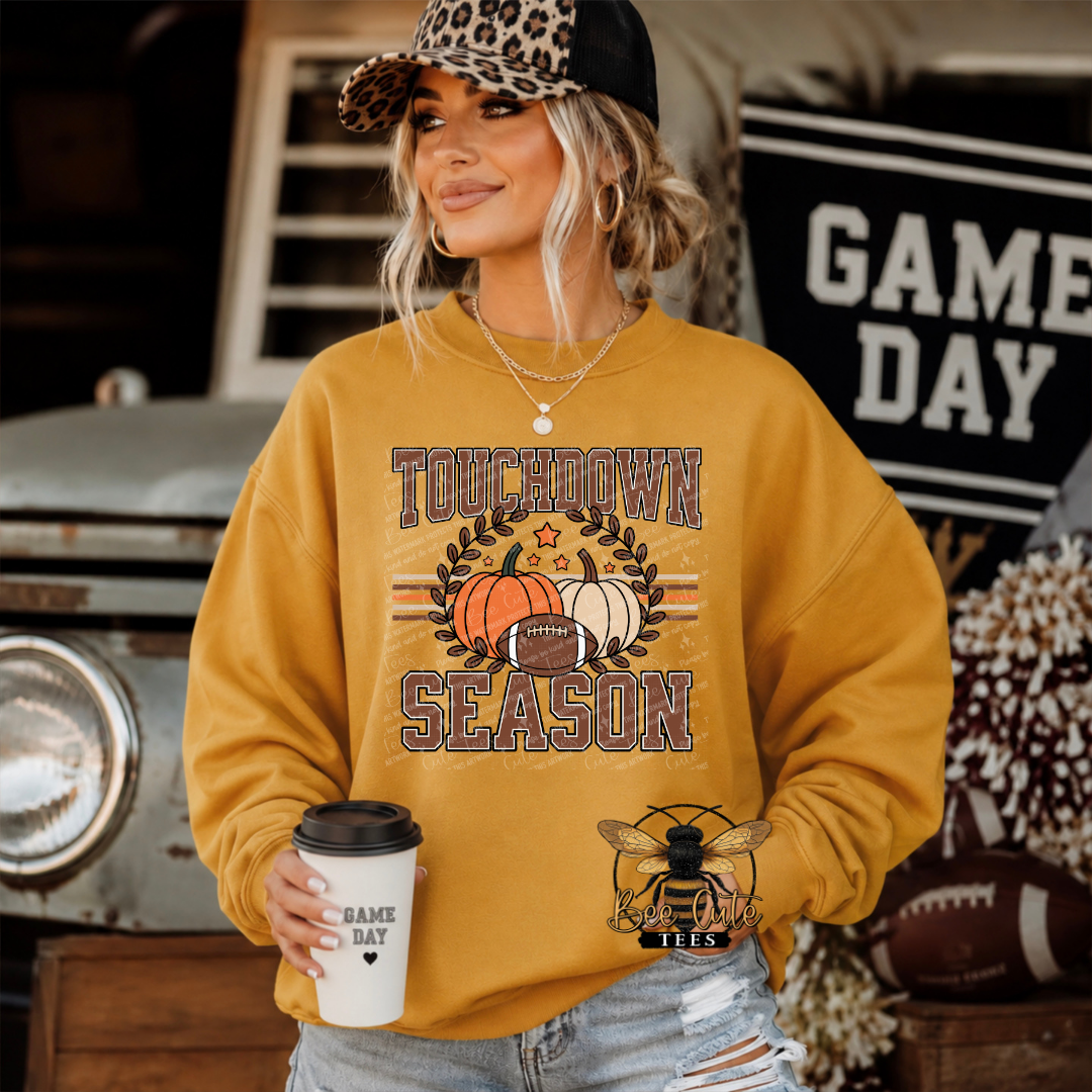 A woman wearing a mustard yellow sweatshirt with a 'Touchdown Season' graphic featuring pumpkins, a football, and a bee logo, holding a coffee cup in a rustic, sports-themed setting.