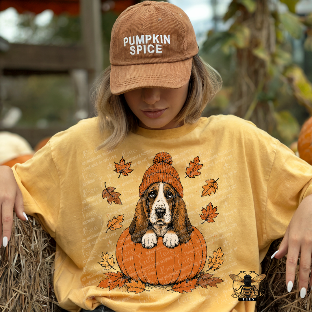 A woman wearing a tan 'Pumpkin Spice' baseball cap and a yellow graphic t-shirt with a basset hound in a pumpkin design, sitting on hay bales at a pumpkin patch.