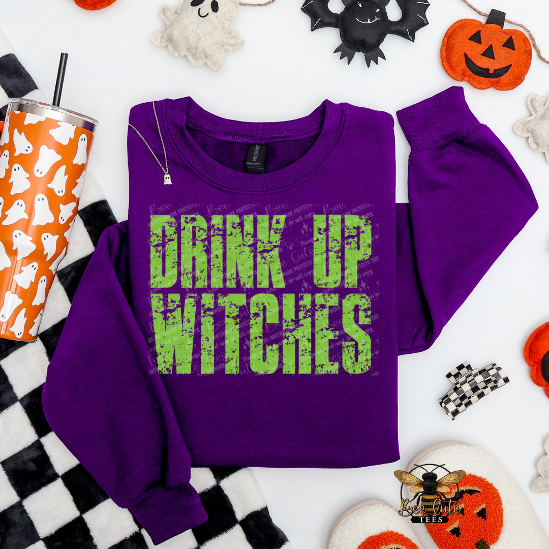 A purple crewneck sweatshirt with the phrase 'DRINK UP WITCHES' printed in distressed green letters, styled in a flat lay with Halloween-themed props including a pumpkin, ghosts, a checkered blanket, and a tumbler.