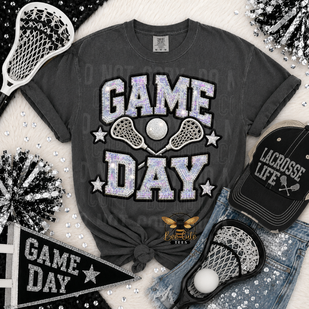 A dark gray graphic t-shirt featuring 'GAME DAY' in glittery silver and lavender letters with lacrosse elements, styled in a flat lay with a black lacrosse cap, denim shorts, a black pennant, and pom-poms on a white faux fur background.
