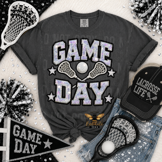 A dark gray graphic t-shirt featuring 'GAME DAY' in glittery silver and lavender letters with lacrosse elements, styled in a flat lay with a black lacrosse cap, denim shorts, a black pennant, and pom-poms on a white faux fur background.