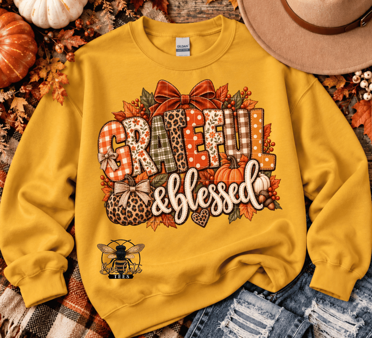 A yellow crewneck sweatshirt laid flat on a rustic autumn-themed background with pumpkins, leaves, denim, and a hat. The front graphic reads “Grateful & Blessed” in mixed patterned letters with bows, leaves, pumpkins, and a bee illustration.