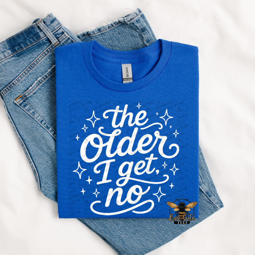A folded royal blue t-shirt with a white graphic reading 'the Older I get, no' surrounded by white stars, styled on top of a pair of blue jeans against a white fabric background.