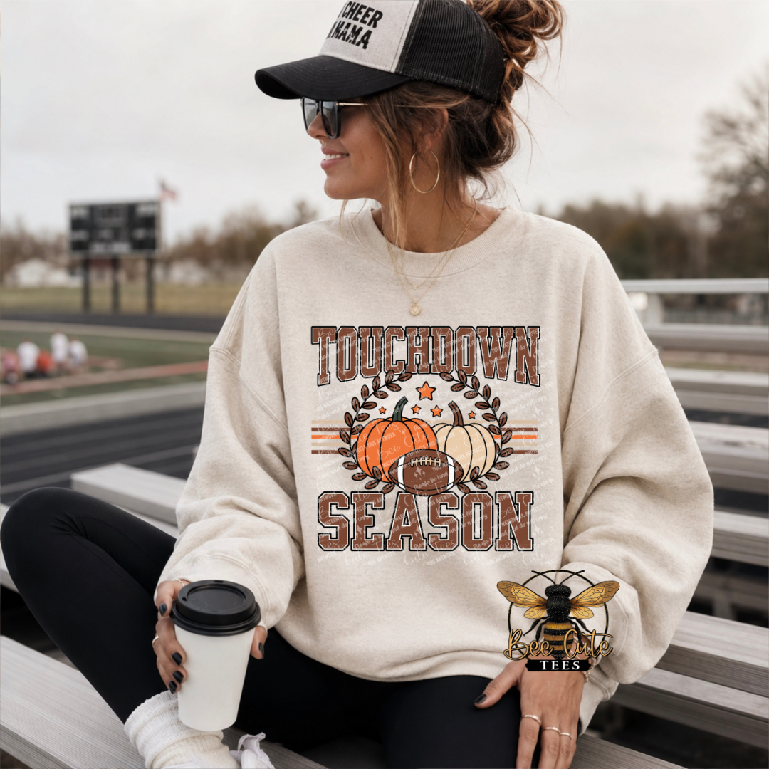 A woman sitting on outdoor bleachers wearing a cream-colored crewneck sweatshirt with a 'TOUCHDOWN SEASON' graphic featuring pumpkins, a football, and a leaf wreath. She is also wearing a black and white trucker hat and holding a coffee cup.