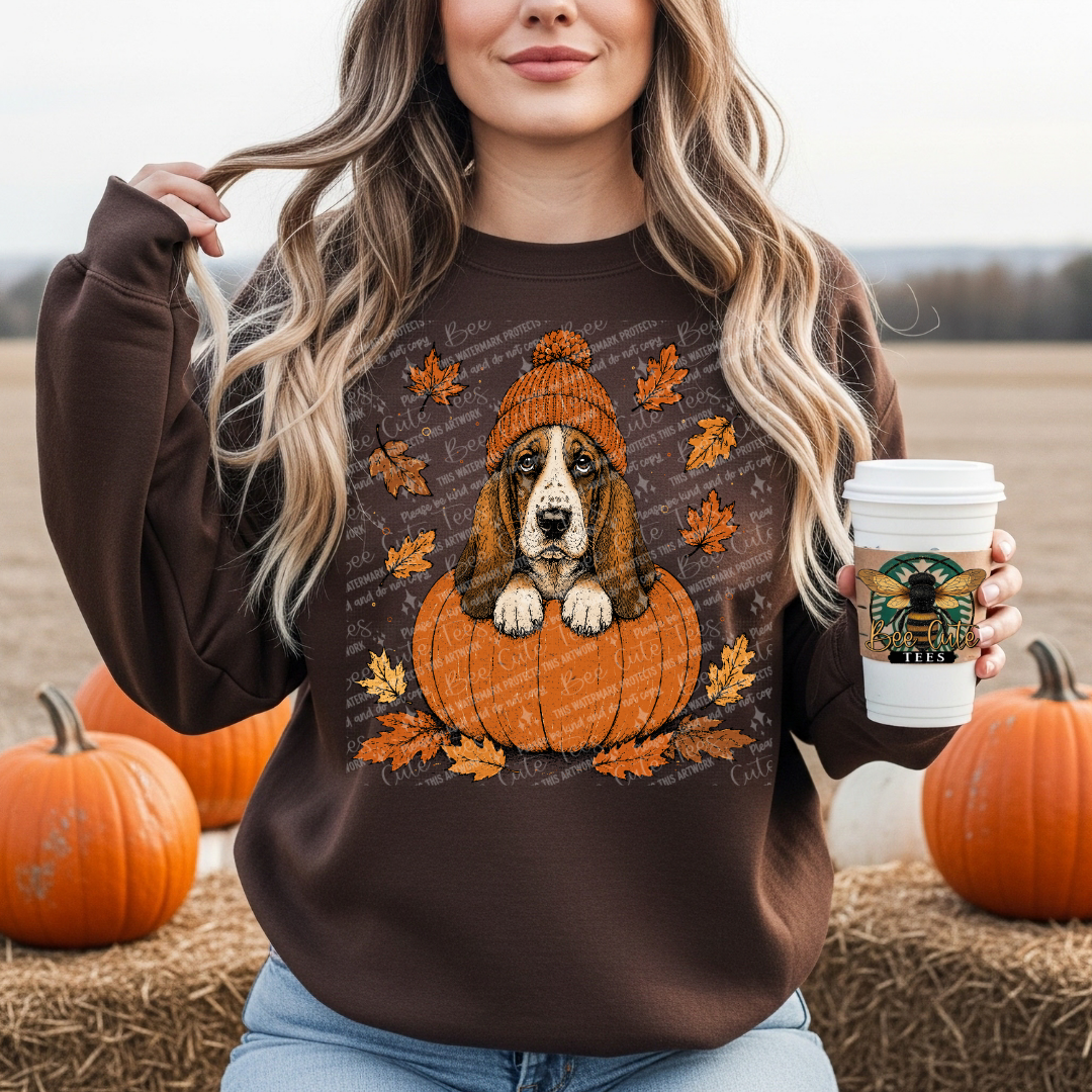 A woman wearing a brown crewneck sweatshirt with a graphic of a basset hound sitting in an orange pumpkin surrounded by autumn leaves. She is holding a white coffee cup and standing outdoors on a hay bale with pumpkins in the background.
