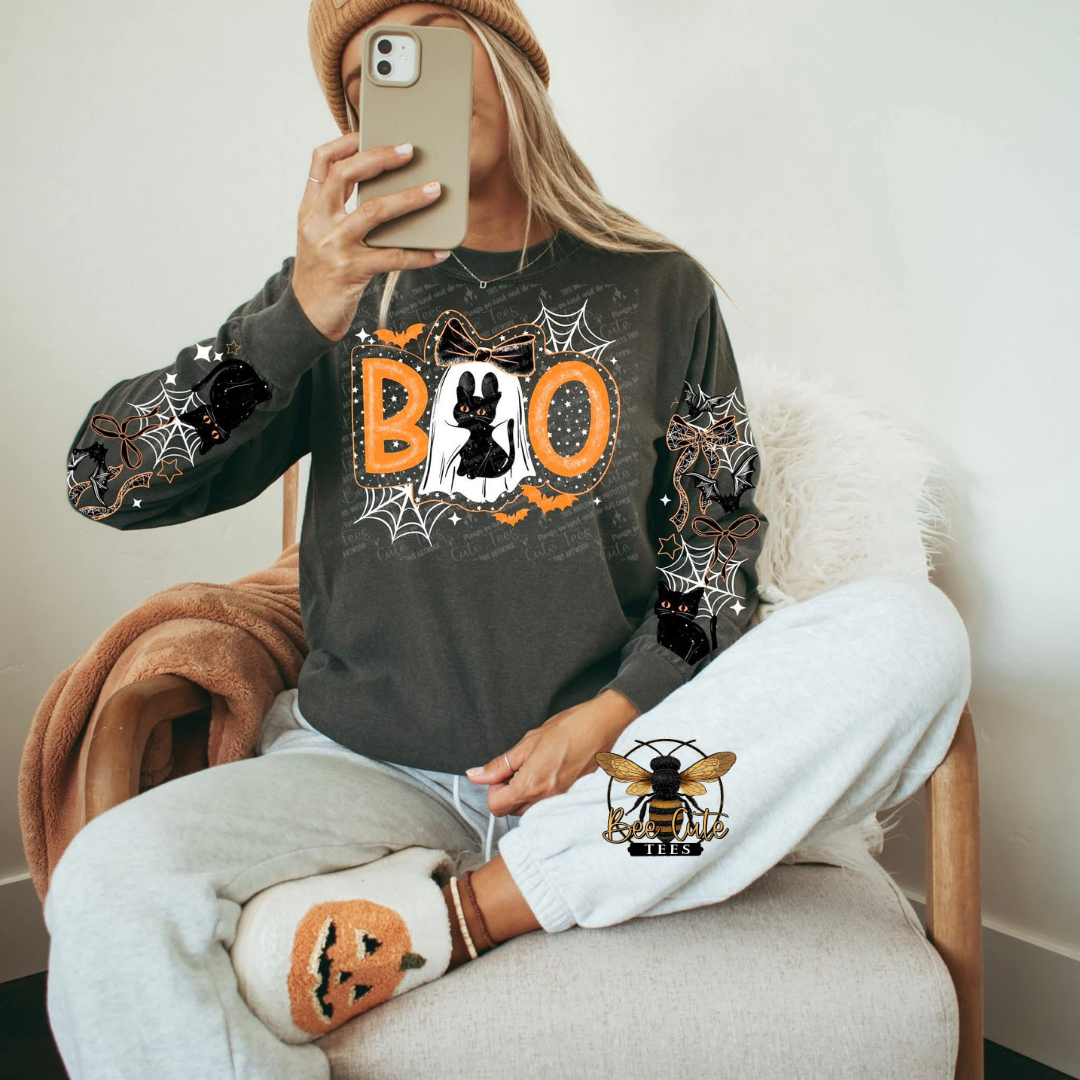 Charcoal gray Halloween sweatshirt featuring a black cat dressed as a ghost inside bold orange “BOO” lettering, surrounded by spiderwebs and bats, with coordinating black cats, bows, stars, and spiderweb designs down both sleeves.
