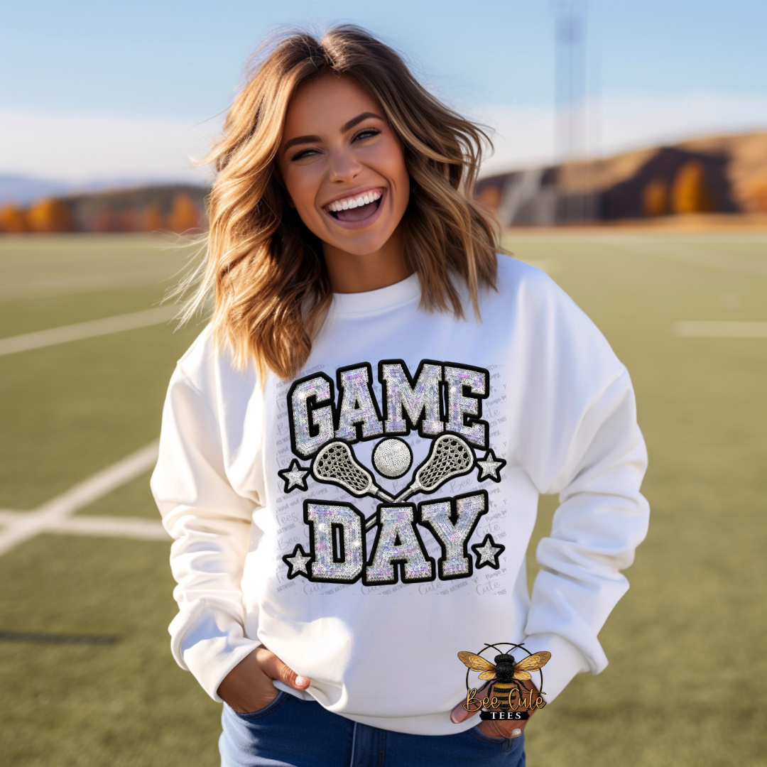A woman smiling on an outdoor sports field wearing a white crewneck sweatshirt with a distressed 'GAME DAY' lacrosse graphic in black and gray, paired with blue jeans.