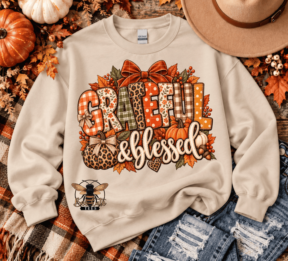 A beige crewneck sweatshirt with a large autumn-themed graphic reading 'Grateful & Blessed' in patterned letters, surrounded by leaves, pumpkins, and a bow. The sweatshirt is styled in a flat lay with a plaid blanket, jeans, a hat, and pumpkins.