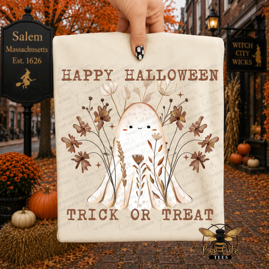 A hand holds a beige folded t-shirt featuring a cute ghost illustration with the words “HAPPY HALLOWEEN” and “TRICK OR TREAT.” The sign is shown against a Halloween-themed street background with autumn leaves, pumpkins, and storefront signs.