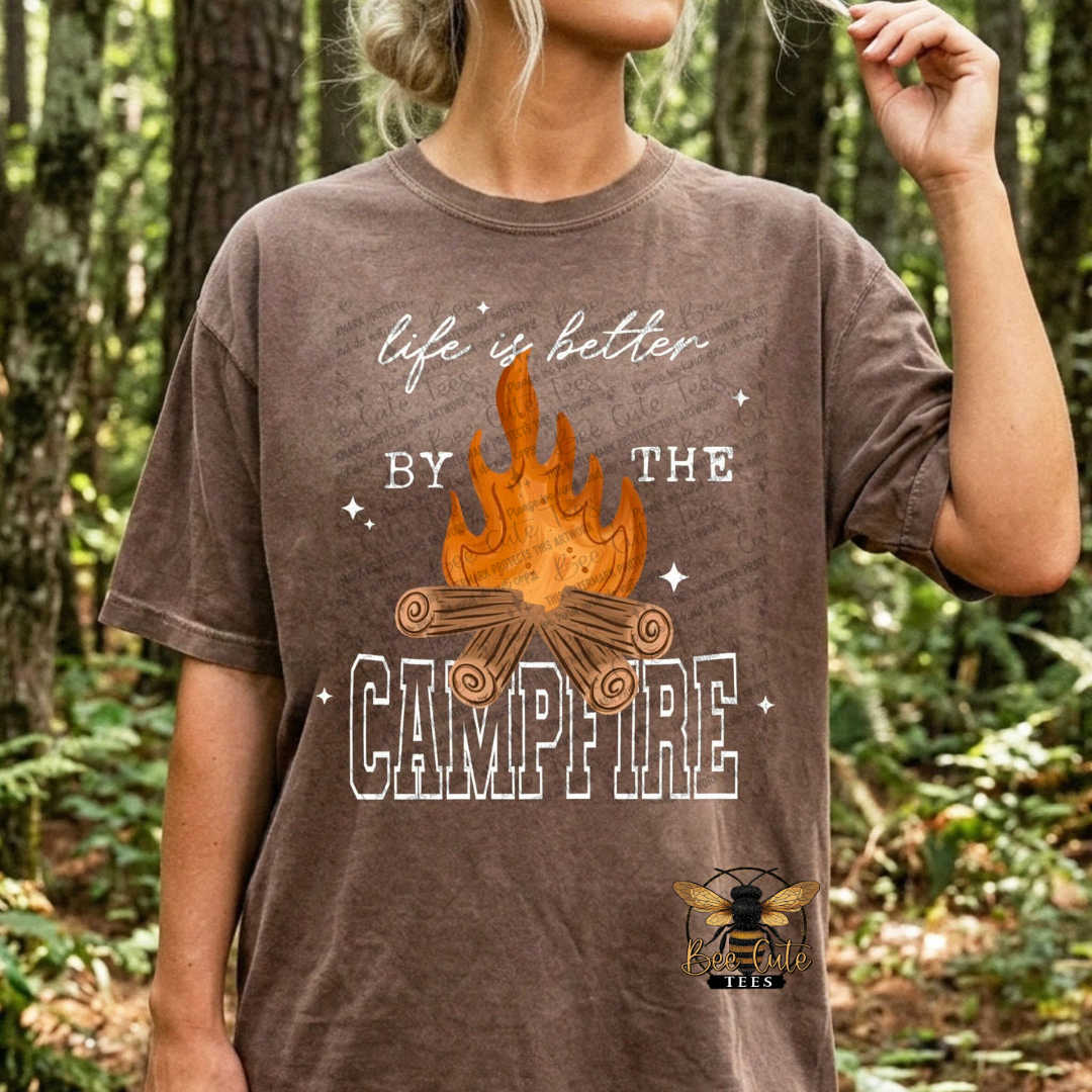 A woman wearing a brown graphic t-shirt in a forest setting. The shirt features a campfire design with the text 'life is better BY THE CAMPFIRE' and a bee logo at the bottom right.