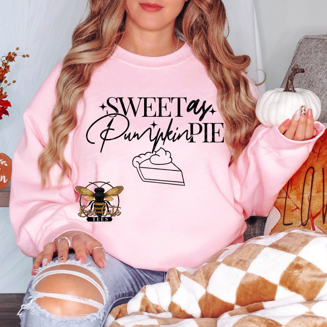A woman wearing a light pink crewneck sweatshirt with a 'Sweet as Pumpkin Pie' graphic and a bee logo on the front, paired with distressed blue jeans.