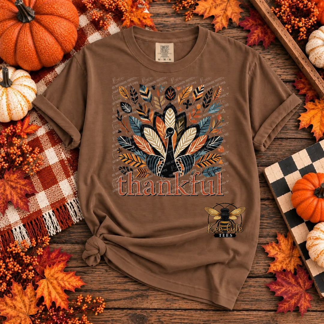 A brown short-sleeve graphic T-shirt laid flat on a rustic wooden surface, surrounded by autumn props including pumpkins, fall leaves, berries, and a plaid blanket. The shirt features a turkey made from layered leaves in orange, beige, blue, and brown, with the word 'thankful' printed below and a bee logo near the lower right front.