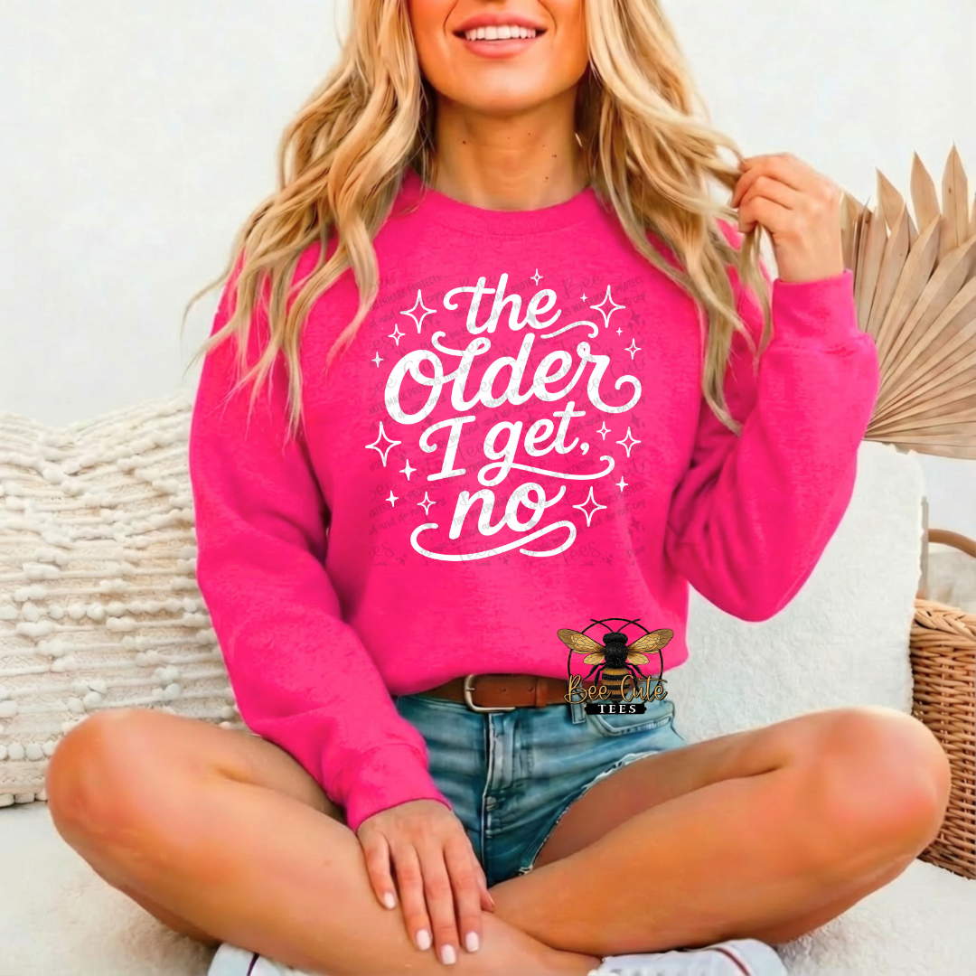 A woman sitting cross-legged on a white couch wearing a bright pink crewneck sweatshirt with the white text 'the Older I get no' and denim shorts.