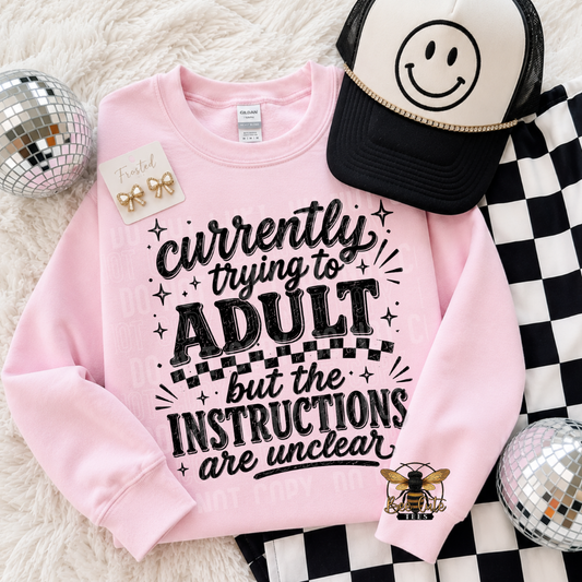A pink crewneck sweatshirt with a black graphic text reading 'currently trying to ADULT but the INSTRUCTIONS are unclear', styled in a flat lay with a black and white smiley face trucker hat, a checkered cloth, and two disco balls on a white faux fur background.